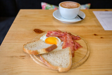 Traditional breakfast meal with fried egg, crispy bacon, slices of bread, and a hot cappuccino served on light wooden table inside a restaurant or cafe, creating a cozy morning scene
