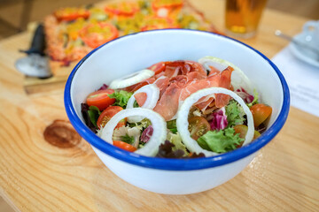 Prosciutto salad mixing fresh green lettuce, cherry tomatoes, and onion rings in a white enamel bowl, on natural wood table with vegan pizza slice on background. Organic and healthy fast food concept.