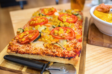 Close up of vegetarian pizza with melted cheese, tomato, zucchini, and corn kernels, being served with cutter on a wooden table on italian restaurant. Healthy and organic fast food concept.