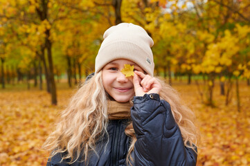 smiling girl in autumn park playfully holds yellow leaf, surrounded by vibrant fall foliage. cozy...