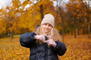 smiling girl in autumn park wearing beanie and jacket forms heart shape with hands. vibrant fall...