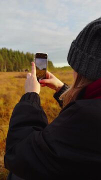 Medium close-up shot from behind of a woman in a grey knitted beanie and black jacket holding a smartphone to capture a wide-angle video of the golden autumn marshland and distant pine trees under 