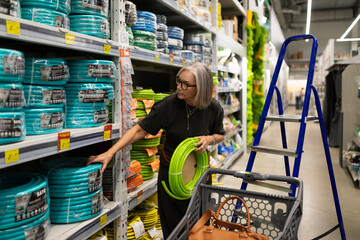 Woman choosing gardening hoses in a hardware store with a shopping cart beside her