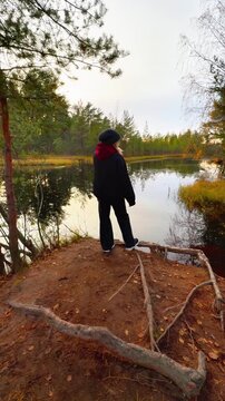 Full shot from a low angle shows a woman wearing a dark jacket and beanie standing on a small, exposed dirt hill overlooking a calm forest lake during a cloudy autumn evening, with exposed tree roots 