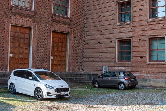 White Ford S-Max and grey Toyota Yaris parked on a cobbled lot in front of classical brick architecture with wooden doors