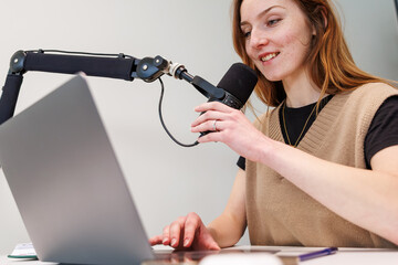 A young woman speaks into a cardioid condenser mic on a boom arm while typing on a silver laptop in a compact home studio. Soft even lighting supports a focused mood.