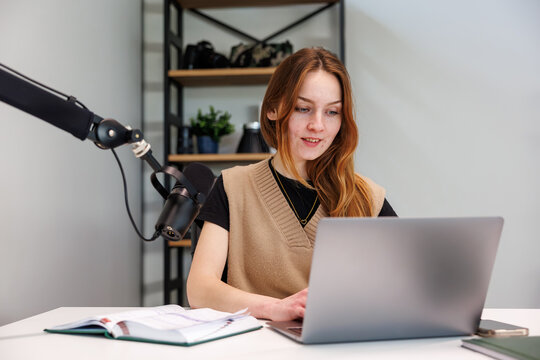 Young woman records at a desk with a laptop and podcast microphone, title in description. She references a notebook and phone as soft daylight and neutral tones set a modern look.