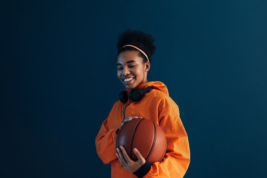 Smiling woman with a basketball wearing orange sportswear and wireless headphones. Young female with curly hair holding a basketball.