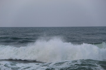 Ocean waves crashing on the shore under a cloudy sky. Blue sea water with white foam shows the power and beauty of nature, perfect for themes of travel, nature, energy, and environment