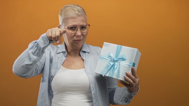 Woman holding a gift box and forming l with hand to forehead in studio orange setting; teasing confidence.