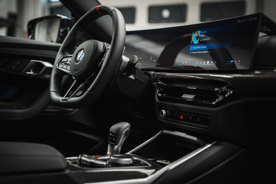 Close interior view shows BMW M2 2025 cockpit from driver side, roundel on tri color stitched wheel, curved display with Welcome, Ravo, carbon trim, compact shifter, soft workshop light