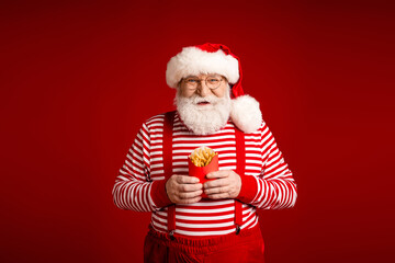 Santa with striped shirt and red suspenders holds a cup of fries against a bright red festive...