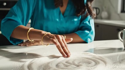 Close-up of a woman's hands drawing designs in a pile of white sugar on a kitchen counter