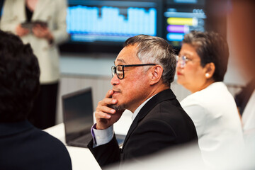 Senior professionals in a modern office meeting, focusing on a thoughtful executive with glasses as colleagues discuss strategy, with data dashboards on screens in the background.
