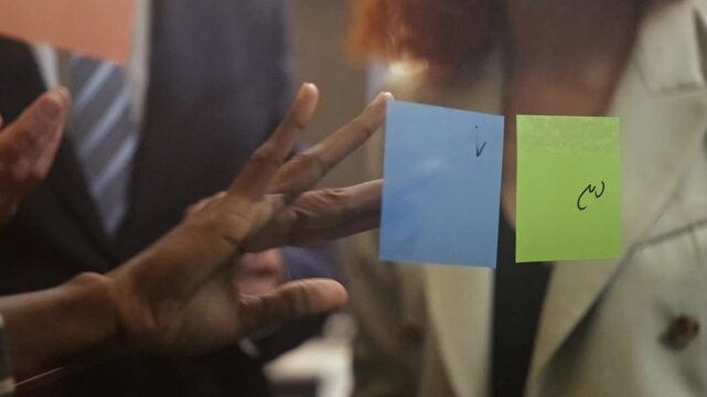Hands placing colorful sticky notes on a glass wall during a business meeting, symbolizing teamwork, planning, and agile project management in a modern office.