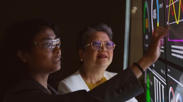 Two professionals analyze data on a large touchscreen, pointing at colorful dashboards and graphs in a dark office, illustrating analytics, teamwork, and decision making.