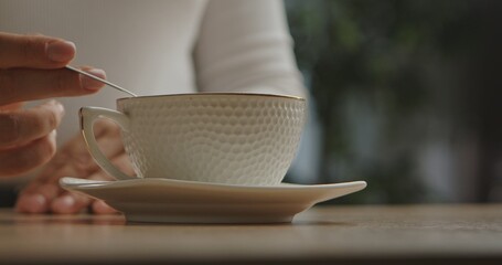 A woman hand gently stirs a hot beverage in an elegant, textured cup placed on a saucer. The scene...