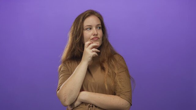 Woman with long hair wearing brown top with hand on chin in studio pensive pose; quiet contemplation.