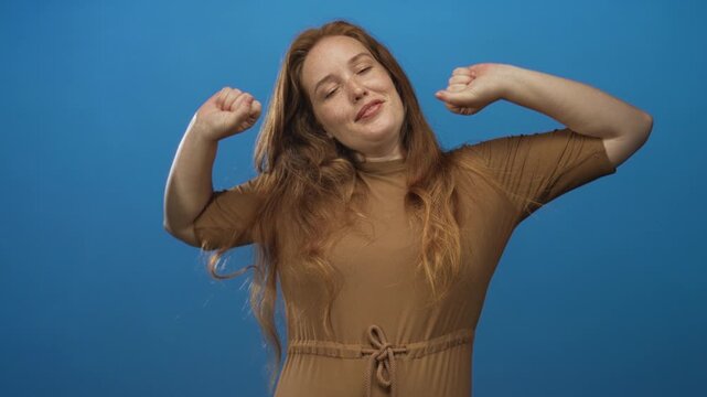 Young caucasian woman yawns while covering mouth with hand in blue studio; fatigue sleepiness drowsiness tiredness.