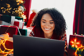 A joyful woman smiles at her laptop surrounded by Christmas gifts and twinkling lights in a cozy...