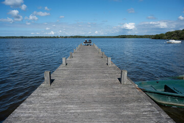Fototapeta premium An elderly couple sits side by side on a bench at Lac de Leon. They enjoy the calm waters and scenic beauty of the Landes region on a clear day.