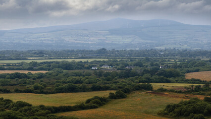 Fototapeta premium Panoramic view of Irish countryside with rolling hills, patchwork fields, trees, and distant wind turbines