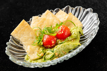A small, clear glass dish contains a dollop of green avocado dip, topped with halved cherry tomatoes, green onions, and sesame seeds, served with crispy triangular crackers.