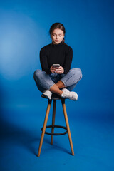 A young woman casually seated on a stool while holding her smartphone against a blue background