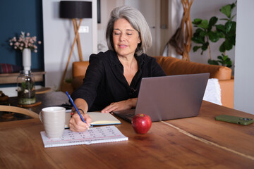 Mature woman writing notes on calendar while working from home using laptop, organizing her schedule