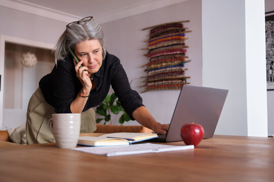 Focused mature businesswoman using laptop and speaking on mobile phone while working remotely from home - Powered by Adobe