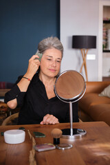 Senior woman massaging her face with a gua sha stone, looking in a mirror on a wooden table at home