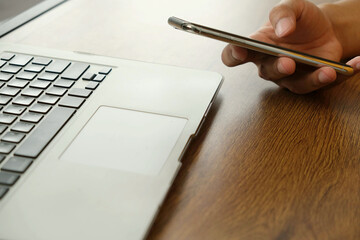 Person using smartphone near laptop on wooden desk, concept of mobile transaction technology and digital communication in modern workspace