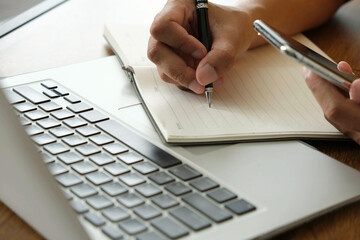 person writing notes in notebook while holding smartphone, with laptop keyboard in view, showing multitasking and business planning