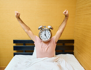 Happy man with alarm clock instead of head stretching on bed at home
