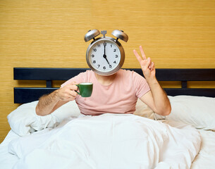 Positive man with alarm clock instead of head holding cup of coffee and showing v sign relaxing in bed