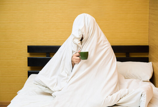 Man wrapping in blanket sitting with cup of coffee in bed