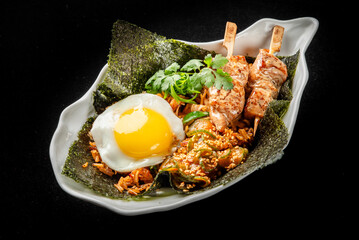A close-up of an Asian-inspired dish served in a white bowl. The meal features fried rice, skewered chicken, a sunny-side-up egg with sesame seeds, cilantro, and nori seaweed.