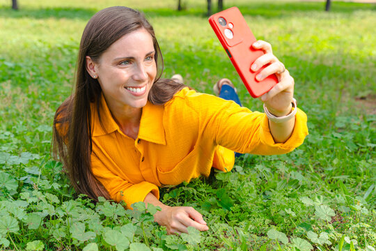Happy woman wearing yellow shirt lying on grass taking selfie with red smartphone in park, enjoying summer day outdoors, social media lifestyle and positive emotions in natural green environment - Powered by Adobe