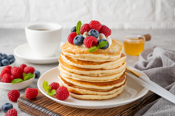 A stack of pancakes with whipped cream, fresh berries and honey on a white plate on a gray concrete background.
