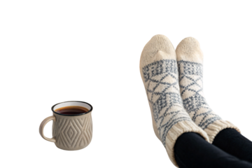 Person's feet in cream knit socks with blue geometric pattern beside textured coffee mug, isolated on a transparent background