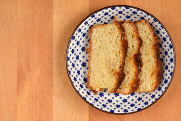A vertical overhead shot of three slices of moist yellow cake with a caramelized top, stacked on a decorative blue and white plate on a light wooden surface