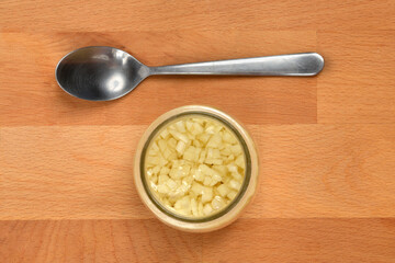 A small glass jar of chopped garlic in oil is centered below a silver tablespoon on a wooden background