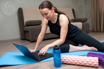 Fit woman following online workout on laptop at home with dumbbells, foam roller and resistance band
