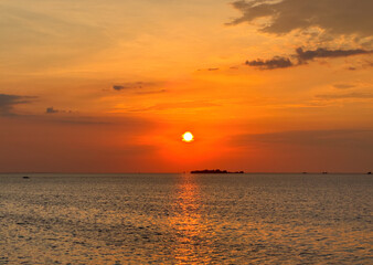 The sun sets over the sea, casting a warm glow across the water and sky. The scene is serene and picturesque, with clouds reflecting the golden hues of the sunset.