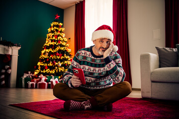 A mature man in festive jumper holding a phone while sitting near decorated Christmas tree in a...