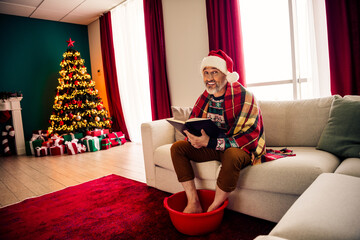 Elderly man enjoying Christmas by the tree while reading indoors, wrapped in a blanket and a cozy...