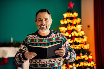 Fototapeta premium Happy senior man in festive sweater reading a book near a Christmas tree at home during holiday time