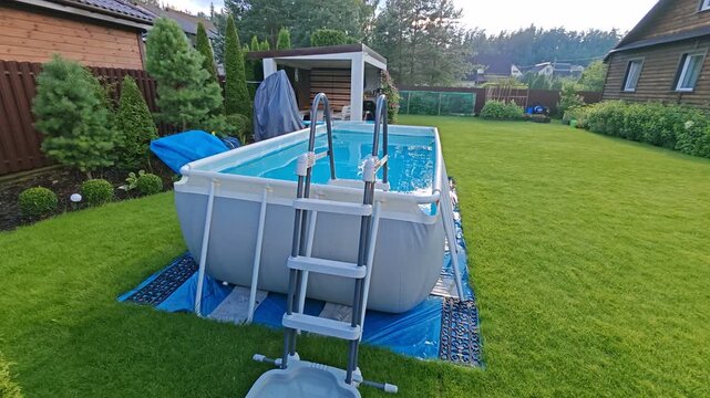 On the mowed grassy lawn stands a gray PVC rectangular frame pool with a ladder. The water in it is filtered. Nearby is a wooden gazebo, swings, house, bushes and trees. Sunny summer weather
