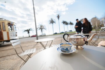 A silver teapot and a delicate blue and white teacup sit on a table outside. A vintage tram and people stroll along the oceanfront in Porto under a bright, cloudy sky