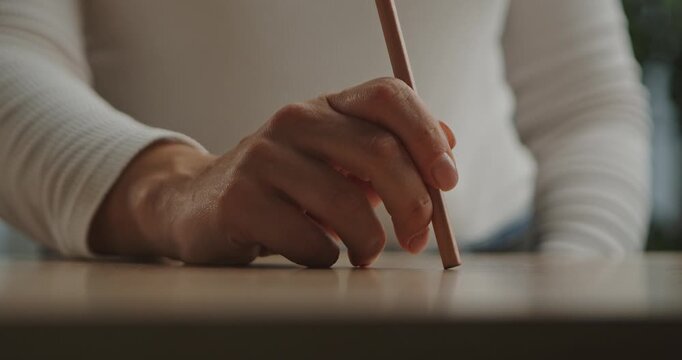 A woman hand taps a wooden pencil on the wooden surface of a table. The room is brightly lit with simple furnishings and a green plant in the background.